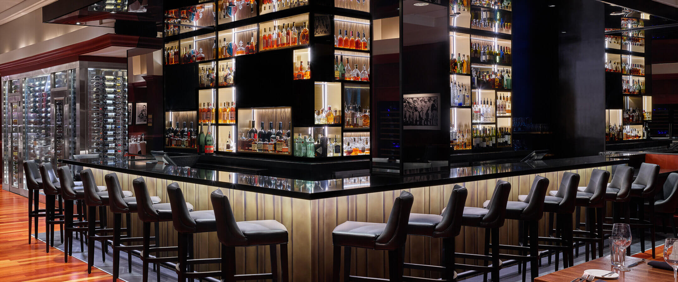 Modern bar with sleek black chairs around a square counter, mirrored shelves filled with bottles, and polished wood floors. The ambiance is upscale and well-lit, with wine racks visible in the background.