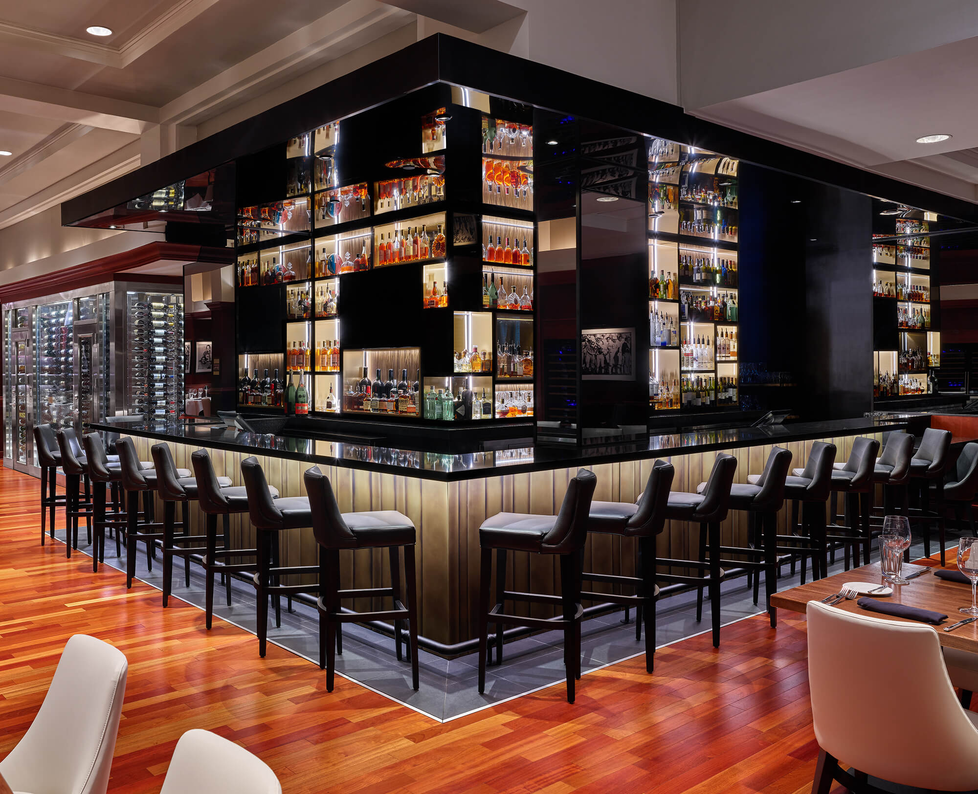 Moderne Bar mit eleganten schwarzen Stühlen um einen quadratischen Tresen, verspiegelten Regalen mit Flaschen und polierten Holzböden. Das Ambiente ist gehoben und gut beleuchtet, mit Weinregalen im Hintergrund.