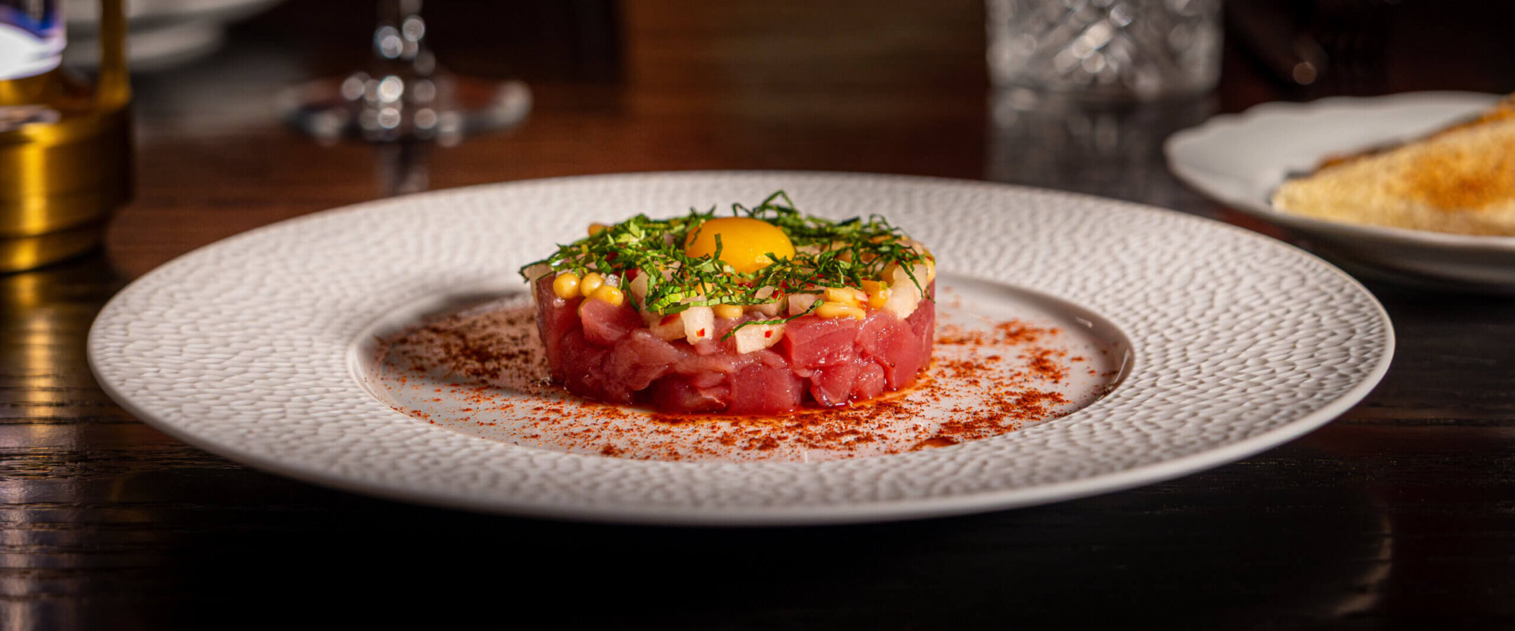 A plate of steak tartare topped with a raw egg yolk and fresh herbs sits on a textured white plate, with a glass of red wine and a water glass in the background.