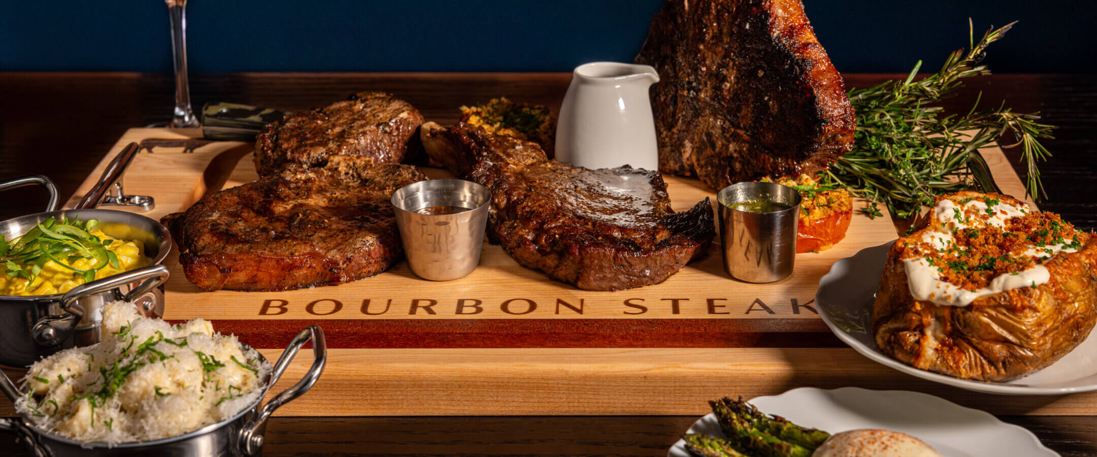 A wooden board labeled Bourbon Steak displays three large grilled steaks with sauces, flanked by dishes of mashed potatoes, white rice, baked potato with toppings, grilled asparagus, and a glass of red wine.