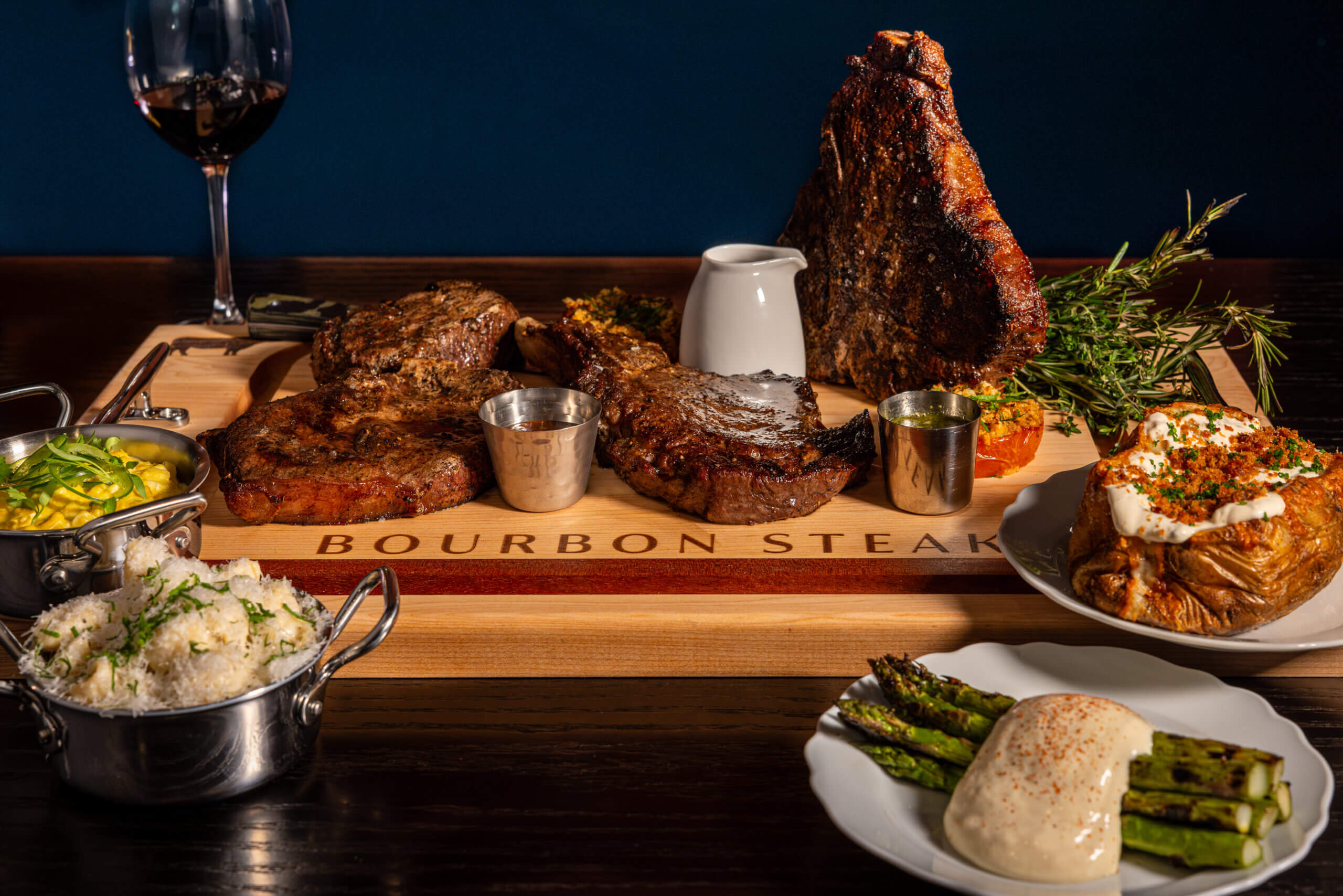 Uma tábua de madeira com o nome Bourbon Steak exibe três grandes bifes grelhados com molhos, ladeados por pratos de purê de batatas, arroz branco, batata assada com cobertura, aspargos grelhados e uma taça de vinho tinto.