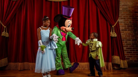 Deux enfants déguisés se tiennent sur scène aux côtés de Mickey Mouse, vêtu d'un costume aux couleurs vives et d'un haut-de-forme. En arrière-plan, on aperçoit des rideaux rouges et un mur de briques. Les enfants sourient et tiennent Mickey par la main.