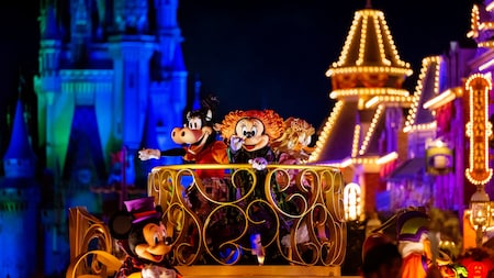 Des personnages Disney vêtus de costumes colorés saluent la foule depuis un char de parade décoré, de nuit, avec en arrière-plan un château et des bâtiments illuminés.