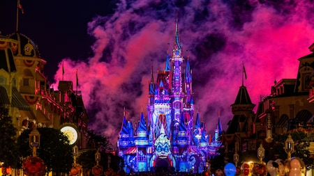 Un château illuminé de lumières rose vif et violettes, avec de la fumée qui s'élève dans le ciel nocturne, entouré de bâtiments se découpant en silhouette et de lampadaires qui brillent.