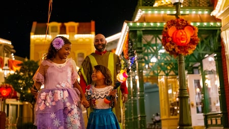 Deux enfants vêtus de costumes colorés et un homme marchent ensemble de nuit dans un quartier festif et très éclairé, décoré de citrouilles d'Halloween et de guirlandes lumineuses. Les enfants sourient et se tiennent par la main.