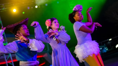 Trois enfants vêtus de costumes colorés exécutent une danse sur scène, sous des lumières d'un jaune et d'un vert éclatants. L'un porte un chapeau de pirate, un autre une robe à fleurs, et le troisième un nœud violet et un tutu.