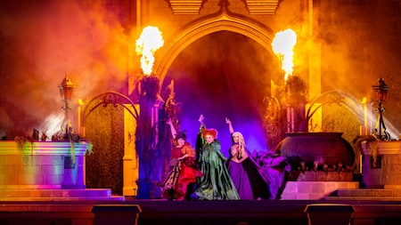 Trois artistes vêtus de costumes colorés se tiennent sur une scène baignée d'un éclairage spectaculaire, ponctuée de jets de feu et agrémentée d'un grand chaudron, créant ainsi une scène magique et théâtrale.