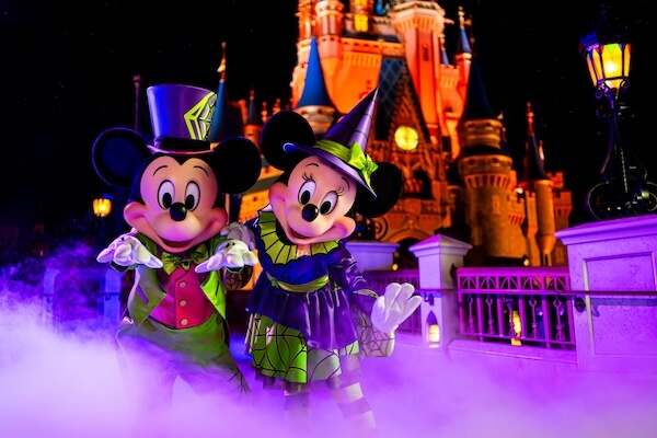 Mickey und Minnie Mouse tragen farbenfrohe Halloween-Kostüme und stehen vor einem hell erleuchteten Schloss in der Nacht, umgeben von lila Nebelschwaden.