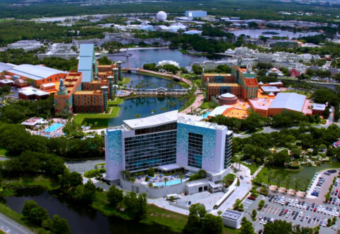 Aerial view of a large resort complex with modern and colorful buildings, pools, and a lake, surrounded by trees, parking lots, and attractions in the background.