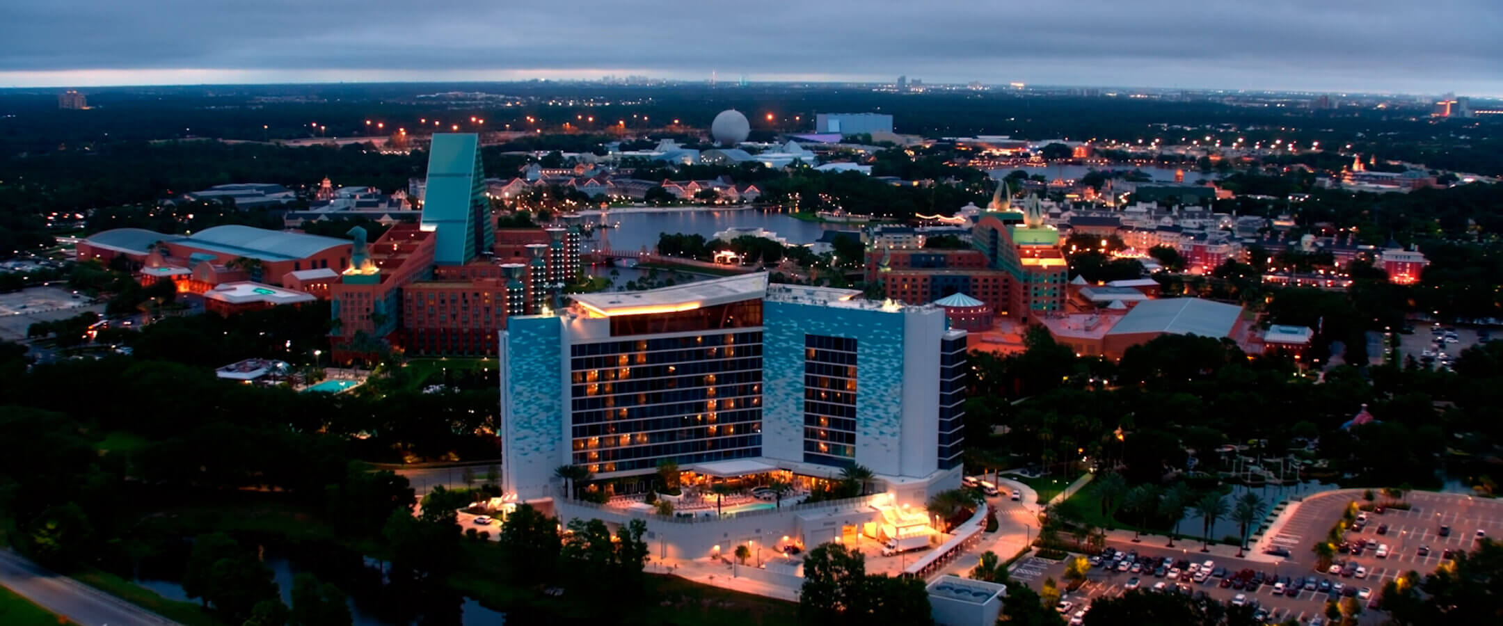 Aerial view at dusk of a modern, illuminated hotel surrounded by trees, with various buildings and attractions in the background and a parking lot in the foreground.