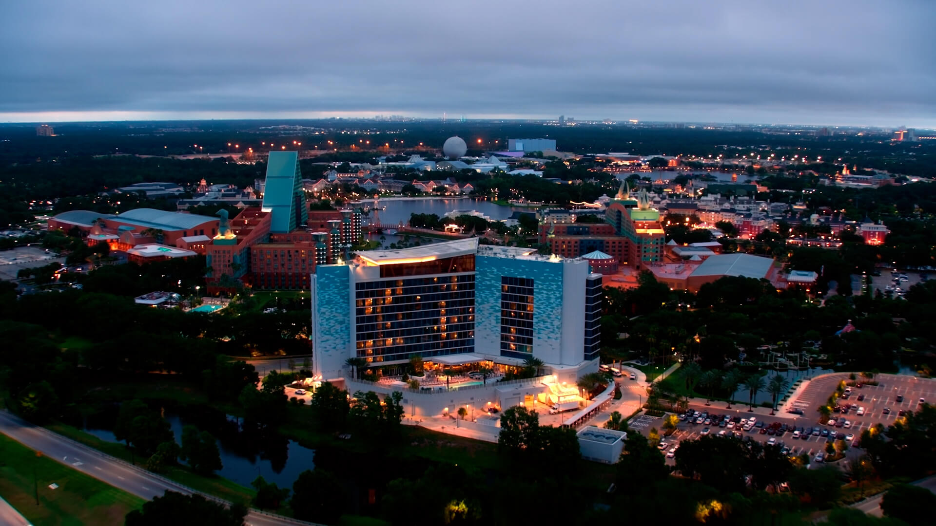 Aerial view at dusk of a modern, illuminated hotel surrounded by trees, with various buildings and attractions in the background and a parking lot in the foreground.