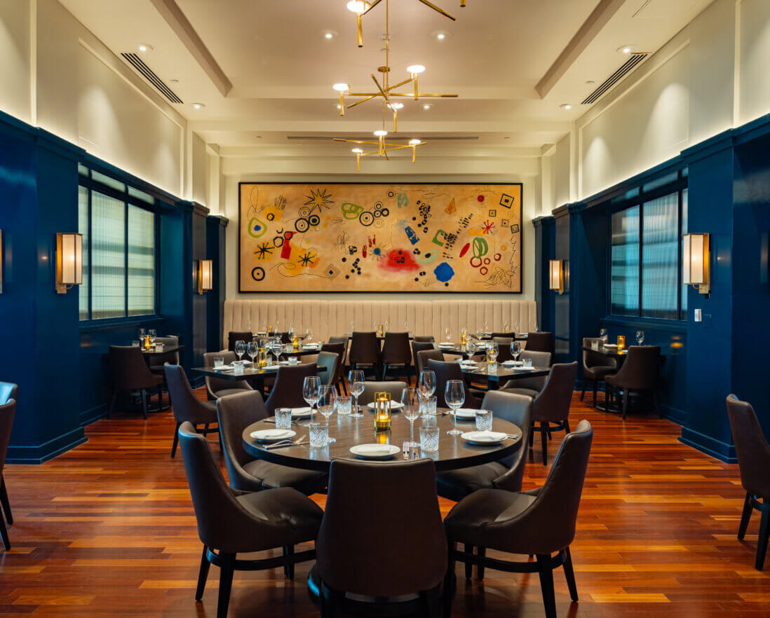 Elegant restaurant dining room with dark blue walls, wooden floors, round tables set with glassware, and modern light fixtures. A large colorful abstract painting is displayed on the back wall.