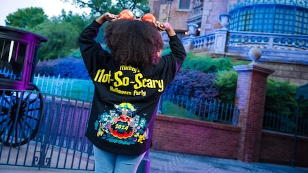 Une personne aux cheveux bouclés, coiffée d'oreilles de Mickey et vêtue d'une veste « Mickeys Not-So-Scary Halloween Party 2024 », se tient devant un bâtiment aux allures effrayantes dans un parc d'attractions.