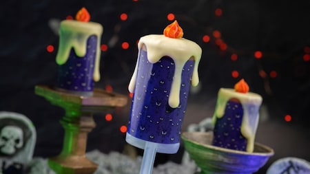 Trois glaces à l'eau violettes décorées pour ressembler à des bougies qui coulent, surmontées de flammes orange, posées sur un fond sombre avec des lumières rouges et des accessoires en forme de pierres tombales.