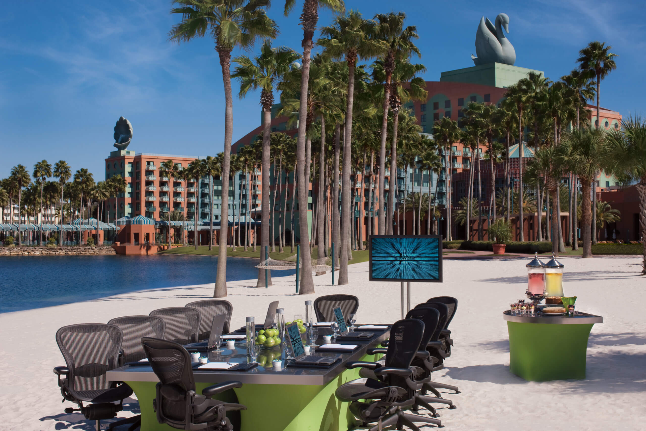 A conference table and chairs are set up on a sandy beach near a lake, with refreshments on a side table, palm trees, and a large hotel building with swan statues in the background.