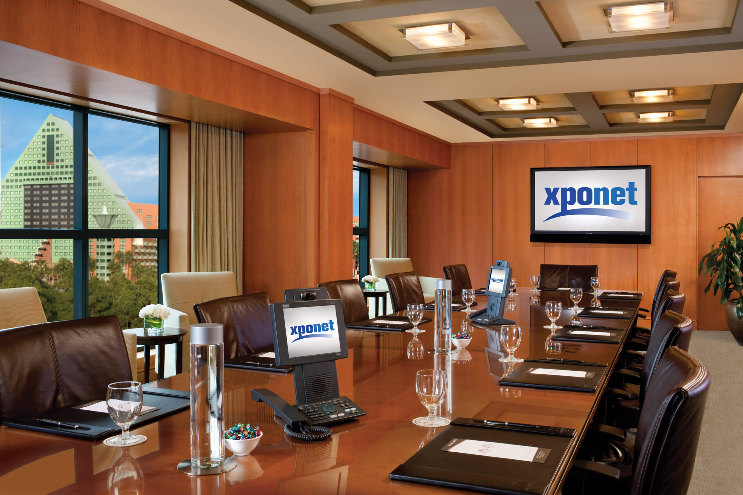 A modern conference room with a long wooden table, leather chairs, digital screens displaying xponet, glassware, notepads, and a window showing a cityscape with a unique pyramid-shaped building.