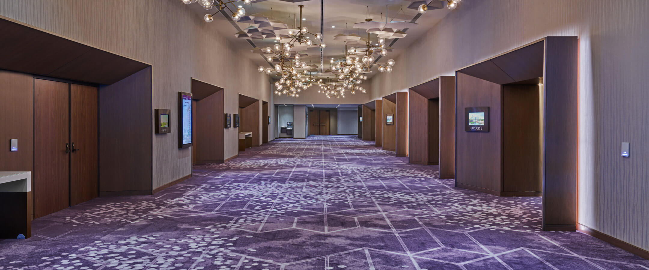 A spacious, modern hallway with purple patterned carpet, geometric ceiling panels, globe chandeliers, and several doorways along the walls. The space is well-lit and elegant, suggesting a hotel or conference center.