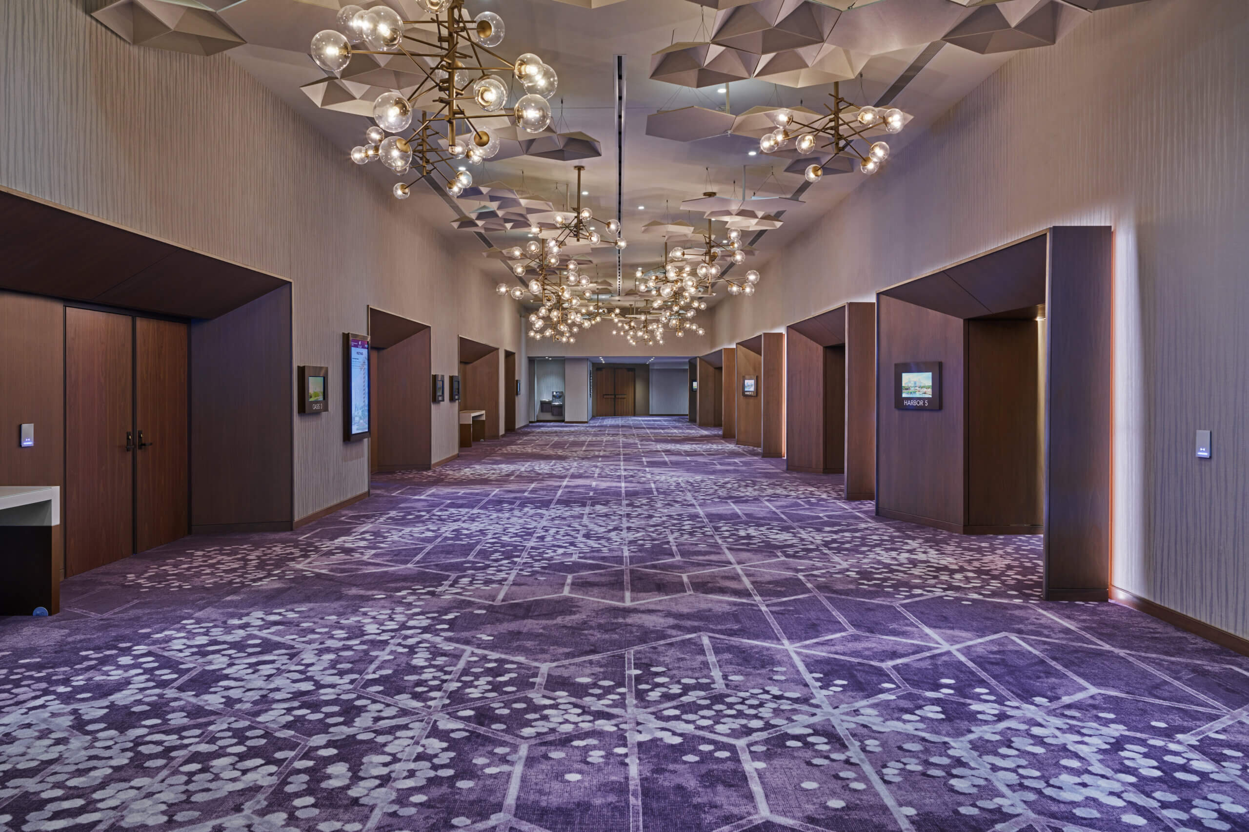 A spacious, modern hallway with purple patterned carpet, geometric ceiling panels, globe chandeliers, and several doorways along the walls. The space is well-lit and elegant, suggesting a hotel or conference center.
