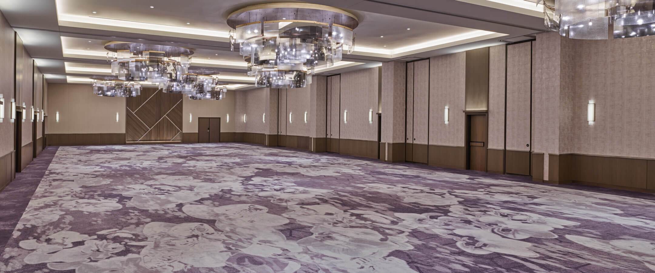 Spacious, modern ballroom with patterned purple floral carpet, neutral walls, large geometric wall accents, and contemporary circular chandeliers hanging from a high ceiling. Room is empty and well-lit.