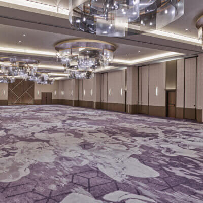 Spacious, modern ballroom with patterned purple floral carpet, neutral walls, large geometric wall accents, and contemporary circular chandeliers hanging from a high ceiling. Room is empty and well-lit.
