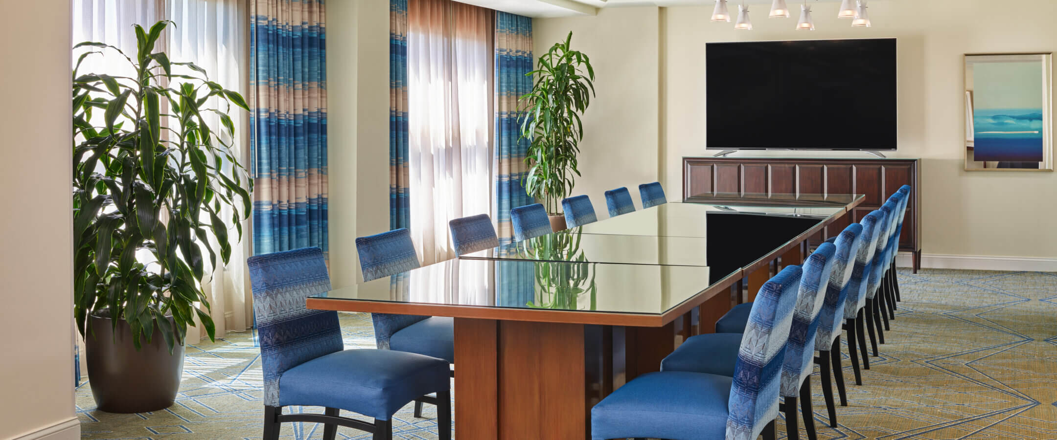 A modern conference room with a long glass table, blue upholstered chairs, large potted plants, a flat-screen TV on a cabinet, patterned carpet, and abstract artwork on the wall. Natural light enters through large windows with sheer curtains.