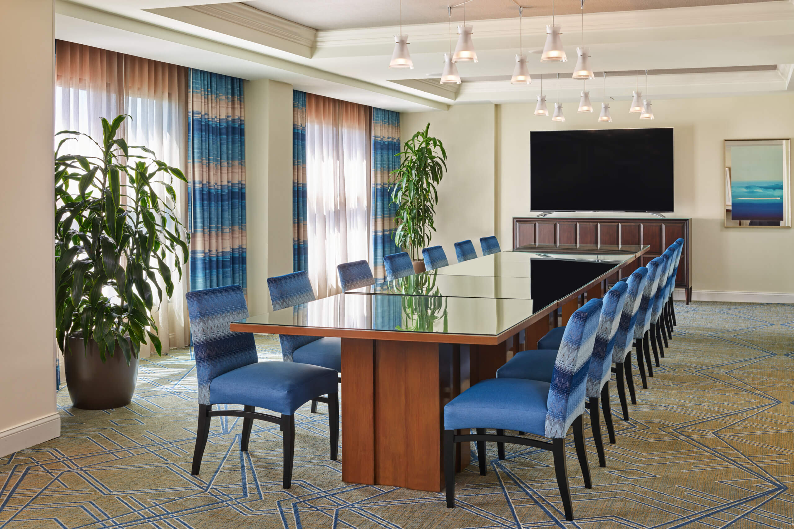 A modern conference room with a long glass table, blue upholstered chairs, large potted plants, a flat-screen TV on a cabinet, patterned carpet, and abstract artwork on the wall. Natural light enters through large windows with sheer curtains.