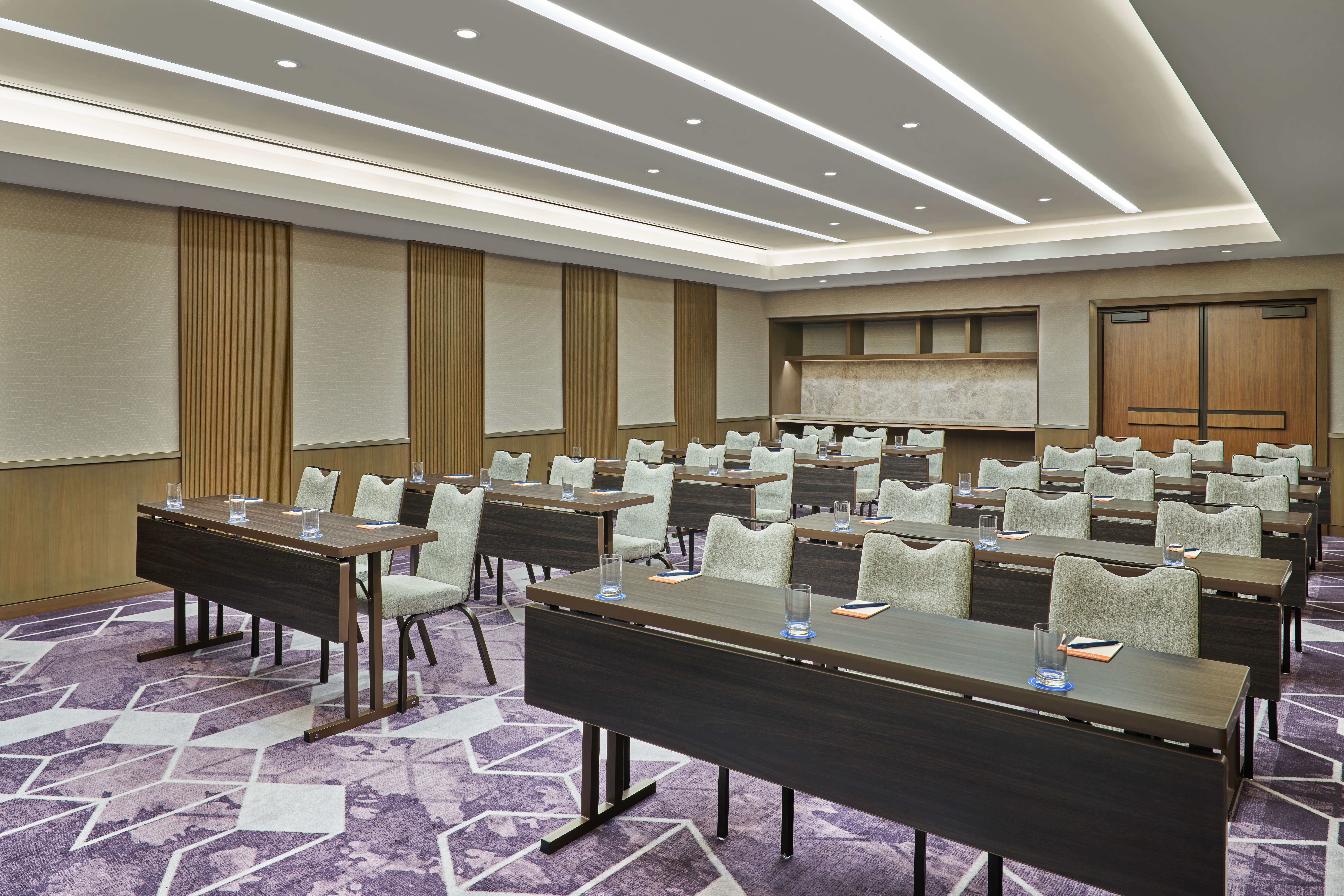 A modern conference room with rows of tables and chairs arranged classroom-style, each table set with a glass of water and notepad. The room features wood paneling, neutral walls, and geometric-patterned purple carpet.