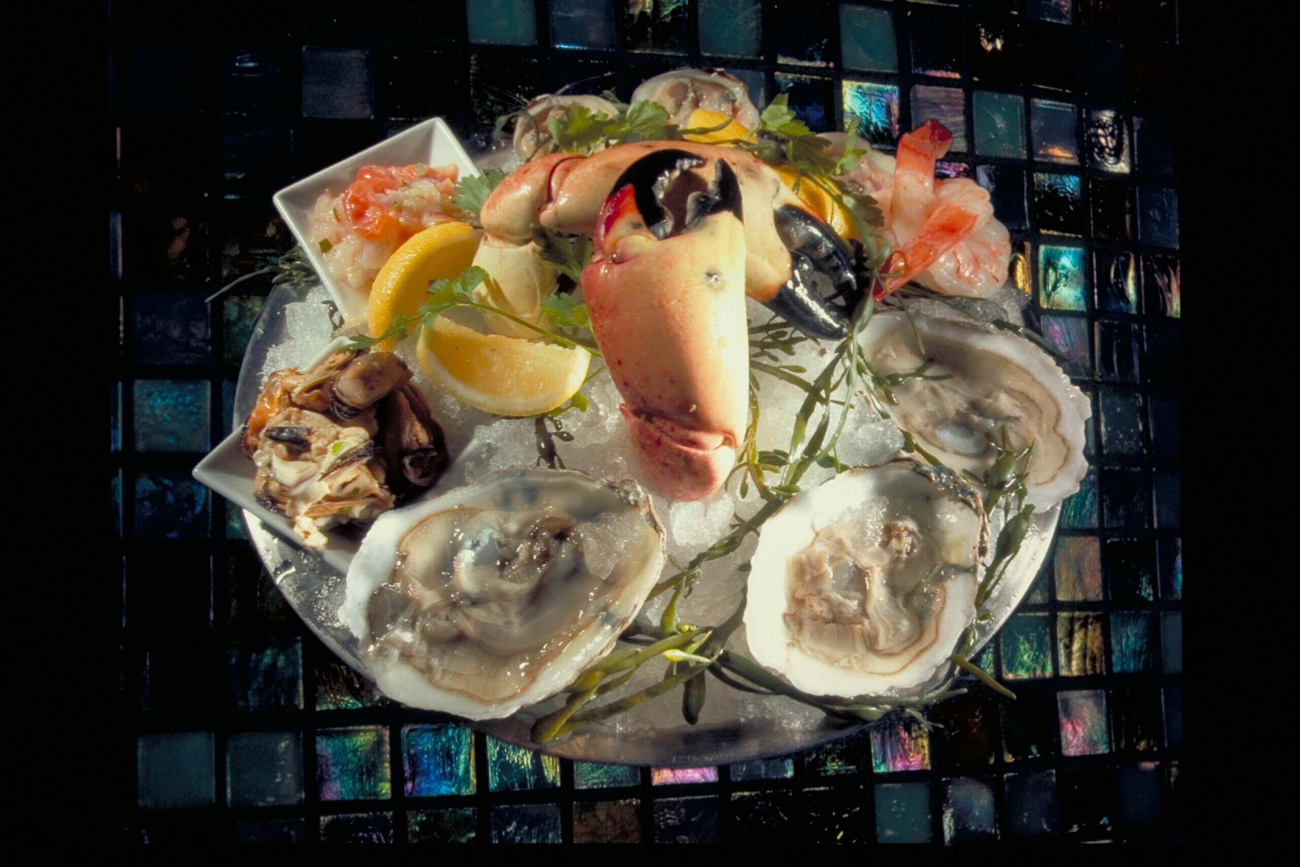 A seafood platter on ice with oysters, crab claws, shrimp, lemon wedges, and garnish, served on a sparkling tiled surface.
