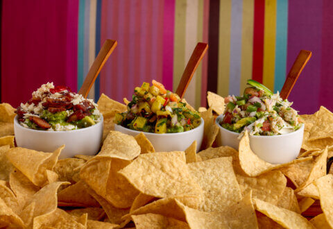 Three bowls of guacamole topped with various ingredients and wooden spoons, surrounded by tortilla chips, with a colorful striped background.
