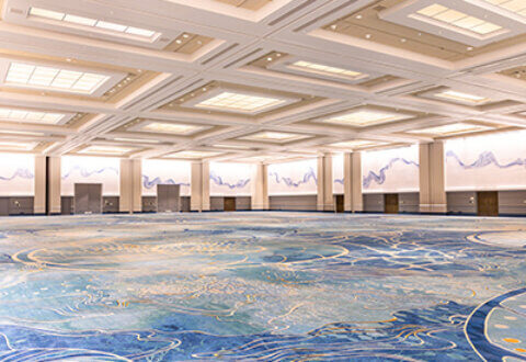 A large, empty convention hall with high ceilings, bright overhead lighting, and a blue patterned carpet. The walls are decorated with abstract blue-and-white designs, and several doors line the perimeter.