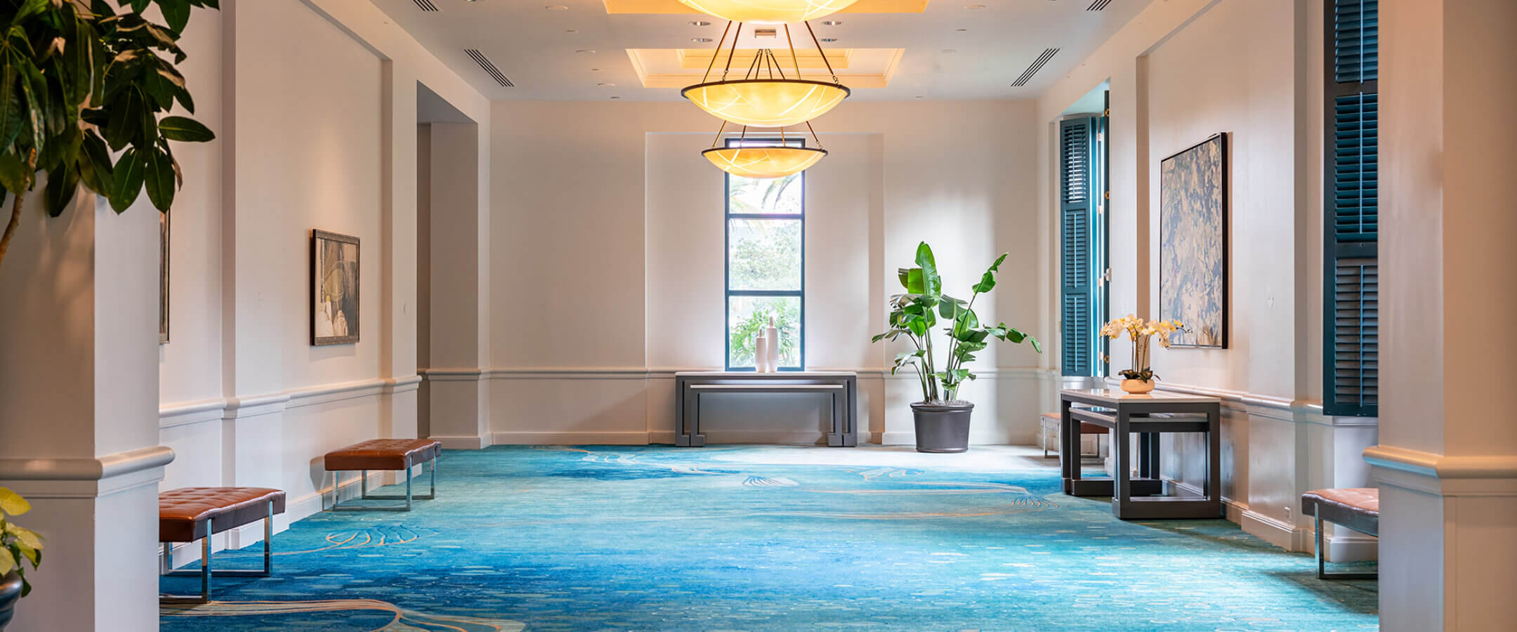 A spacious hallway with blue patterned carpet, modern chandeliers, benches along the walls, artwork, plants, and a window at the far end letting in natural light.