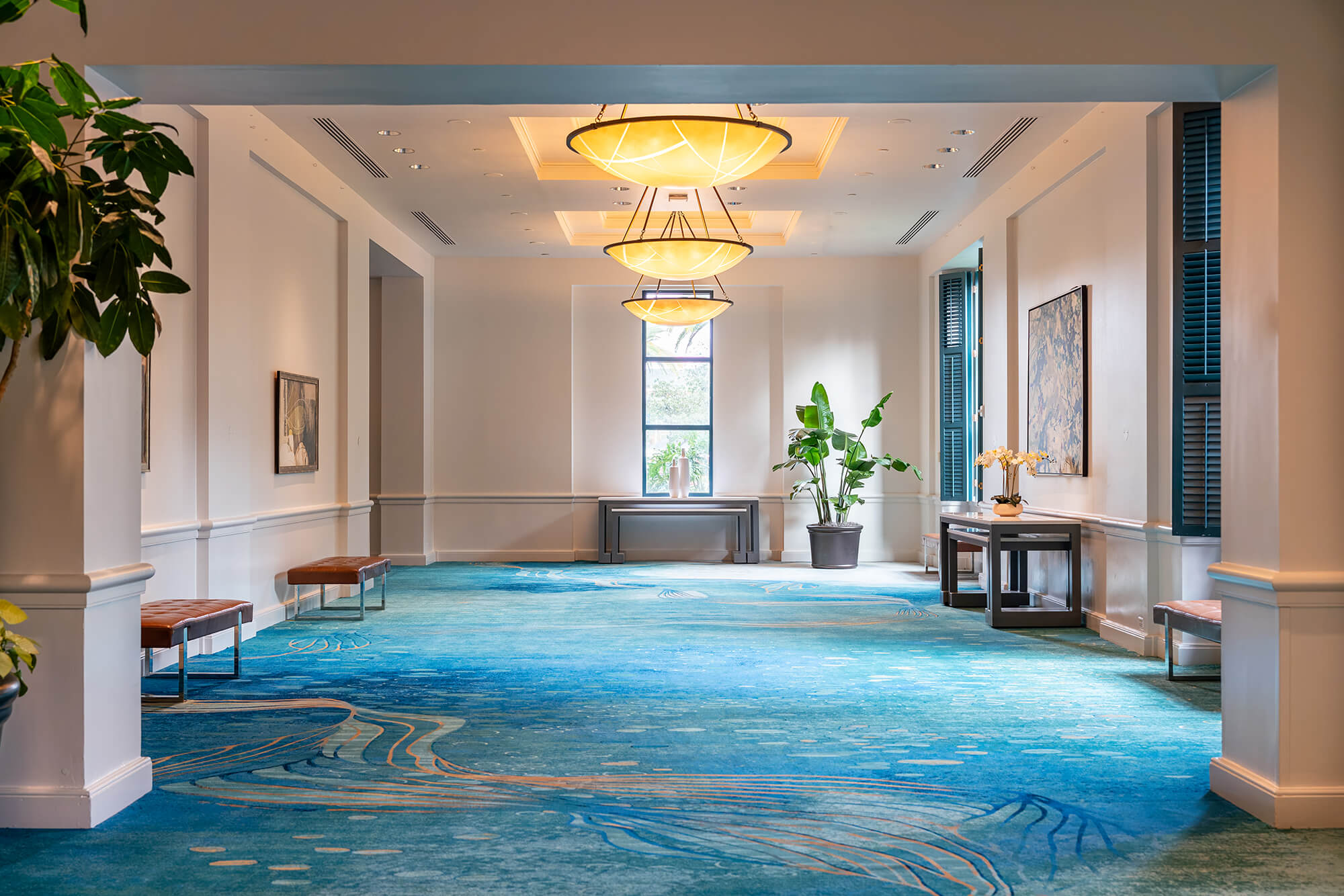 A spacious hallway with blue patterned carpet, modern chandeliers, benches along the walls, artwork, plants, and a window at the far end letting in natural light.