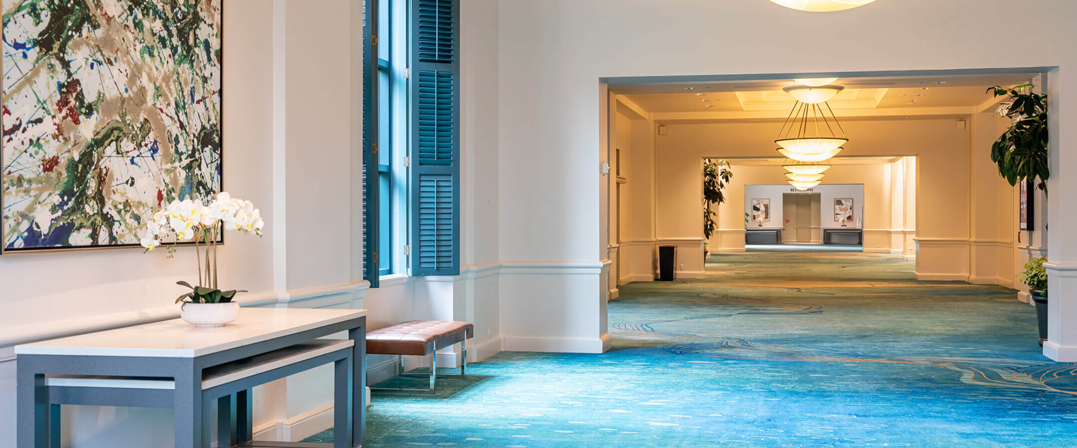 A brightly lit hallway with blue patterned carpet, modern light fixtures, large abstract artwork on the wall, and a table with a white orchid. Windows and a bench are on the left, leading to another well-lit corridor.