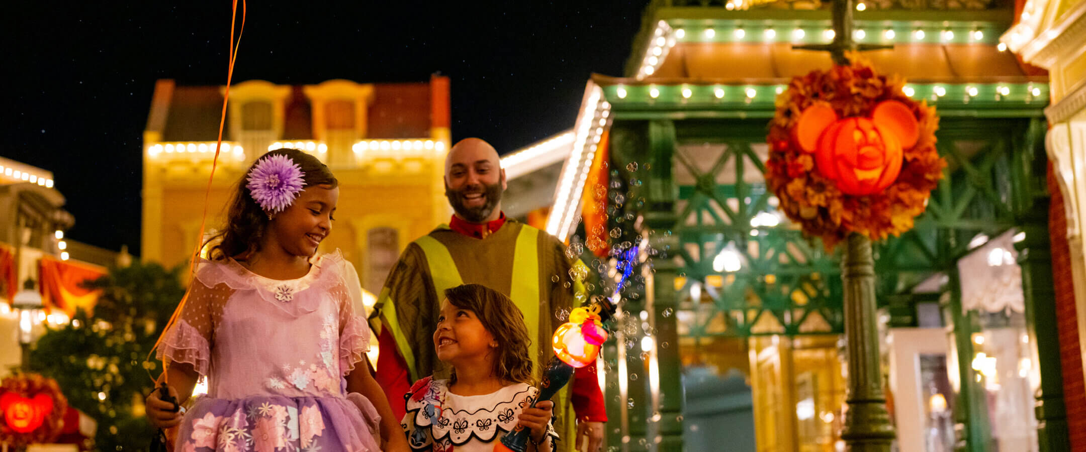 Two children in colorful costumes and a smiling adult walk together at night in a festively decorated street with Halloween lights, wreaths, and a balloon that says Happy Halloween.