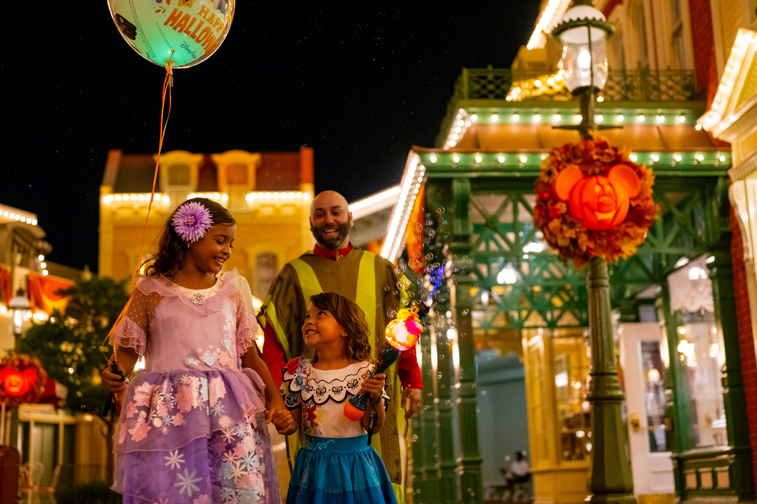 Deux enfants en costumes colorés et un adulte souriant marchent ensemble la nuit dans une rue décorée de lumières d'Halloween, de couronnes et d'un ballon qui dit "Joyeux Halloween".