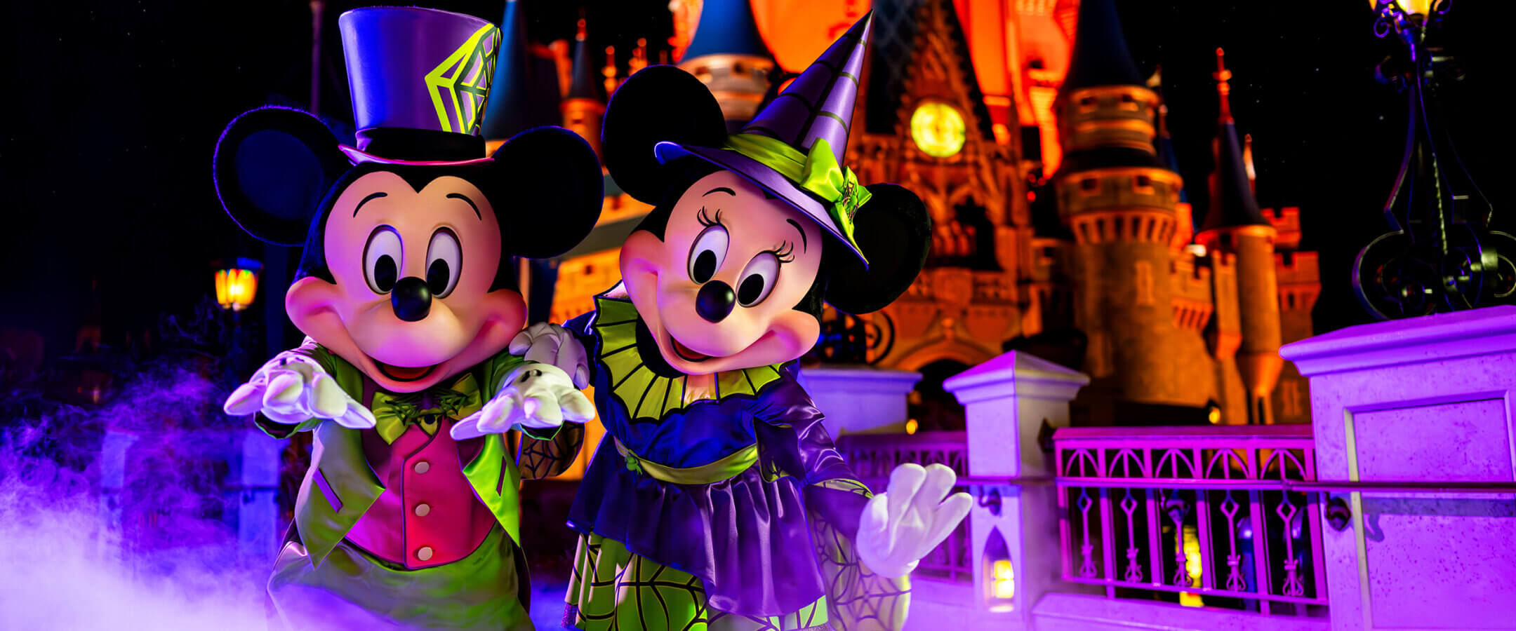 Mickey and Minnie Mouse dressed in Halloween costumes, standing in front of a brightly lit castle at night, surrounded by purple fog.