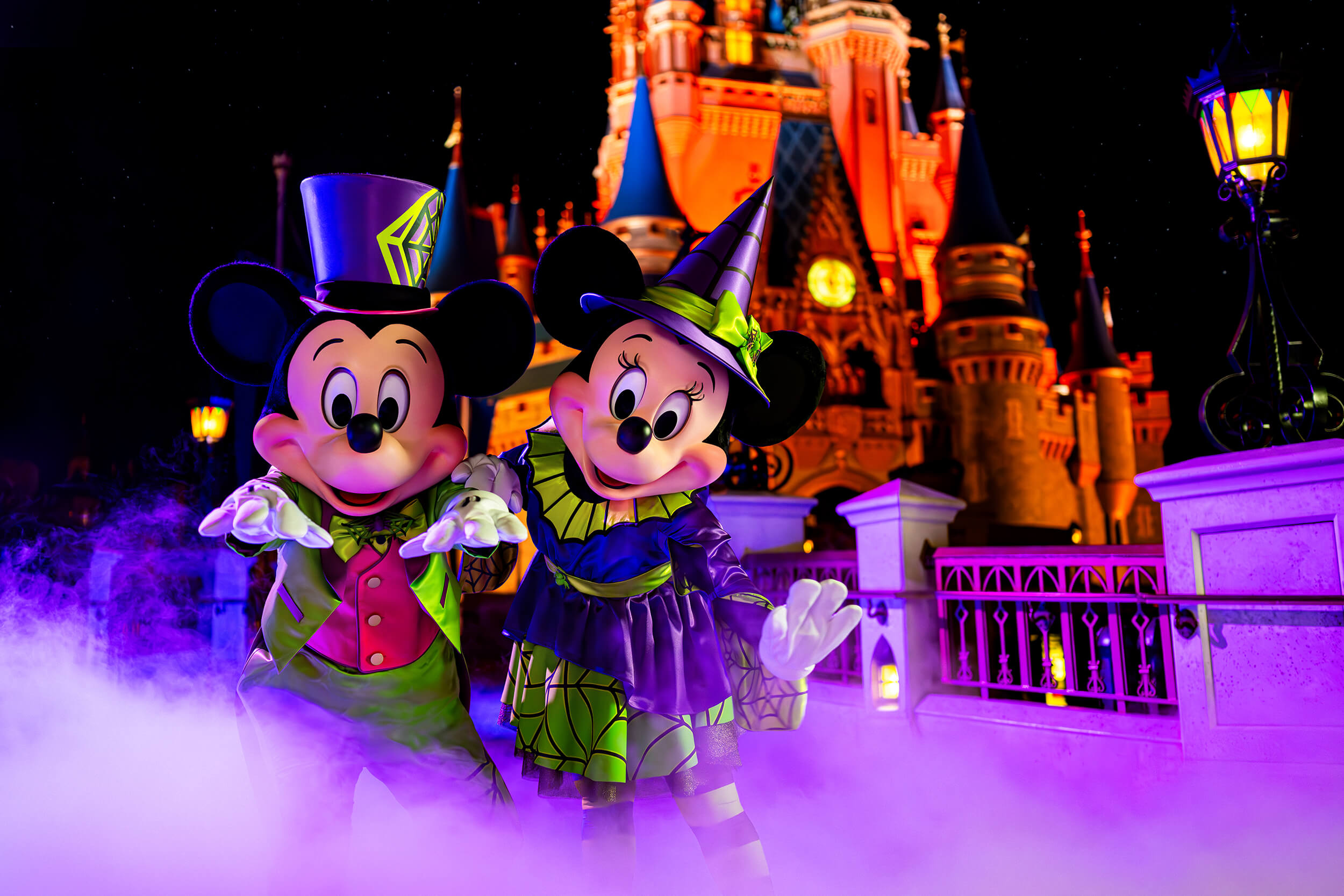 Mickey et Minnie Mouse vêtus de costumes d'Halloween, debout devant un château illuminé de nuit, entouré d'un brouillard violet.