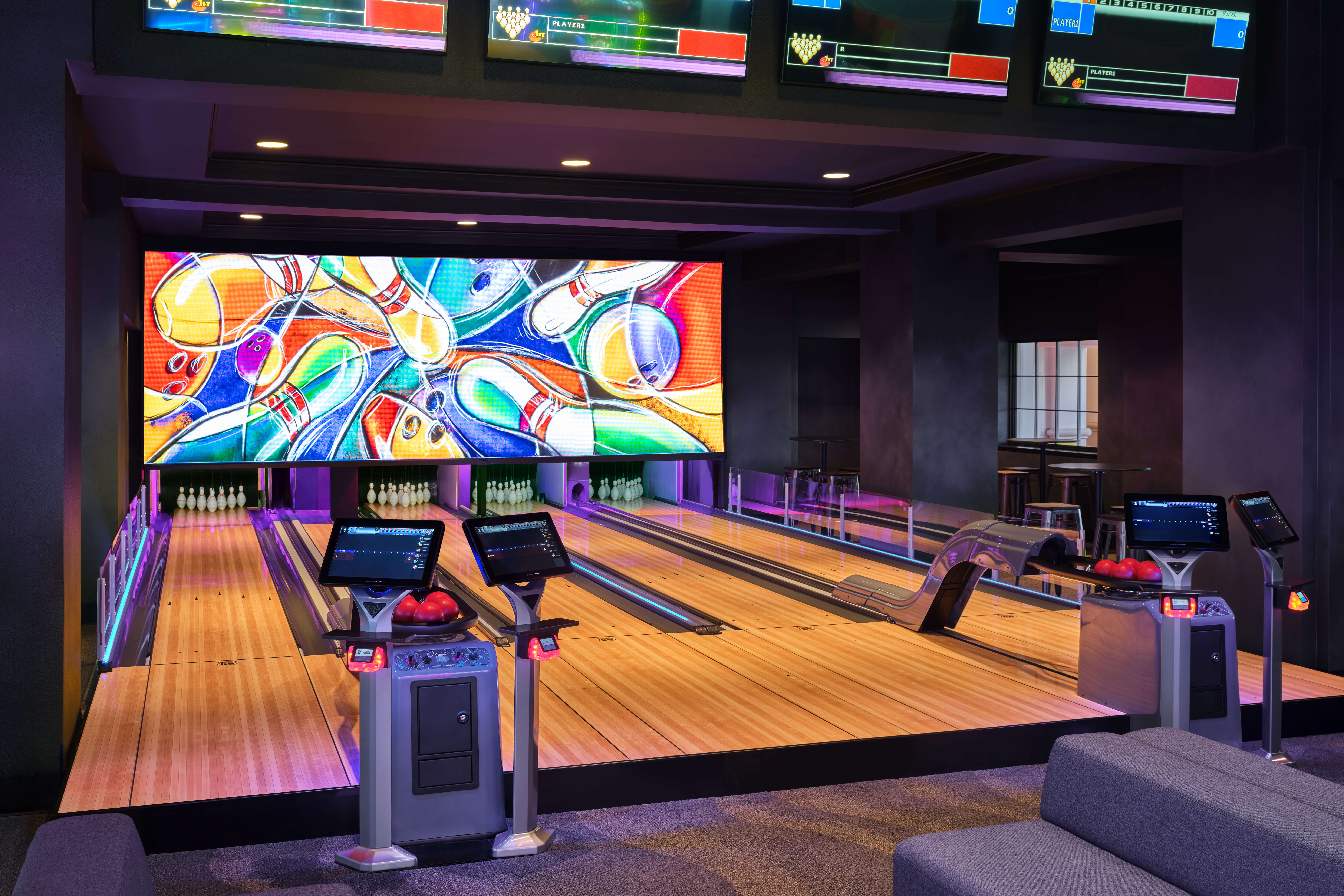 A modern bowling alley with two lanes, electronic scoring screens, bowling balls, and a colorful mural of pins and balls above the pins. The area is well-lit and features sleek, contemporary design elements.