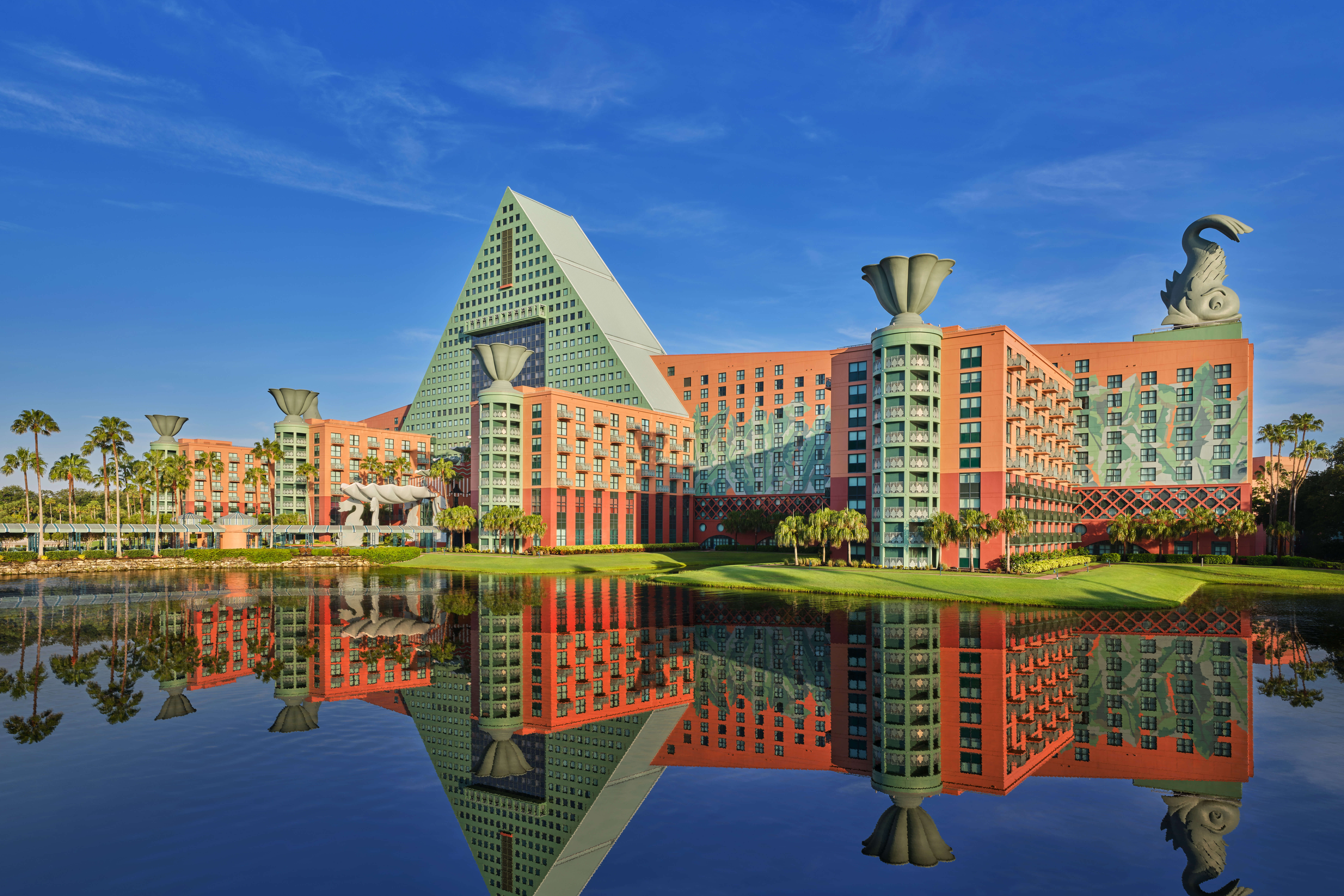 A large, colorful hotel with a pyramid-shaped central tower and whimsical rooftop sculptures is reflected in a calm lake under a clear blue sky, surrounded by palm trees and green lawns.