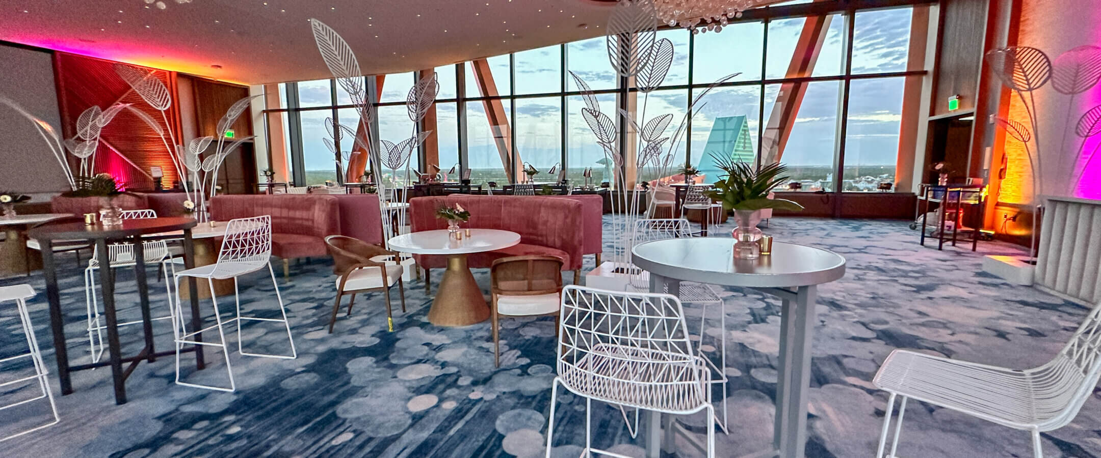 A modern, stylish lounge with blue patterned carpet, white chairs and tables, and large windows offering a city view. Ceiling features artistic light fixtures, and walls are lit with purple and orange lights.