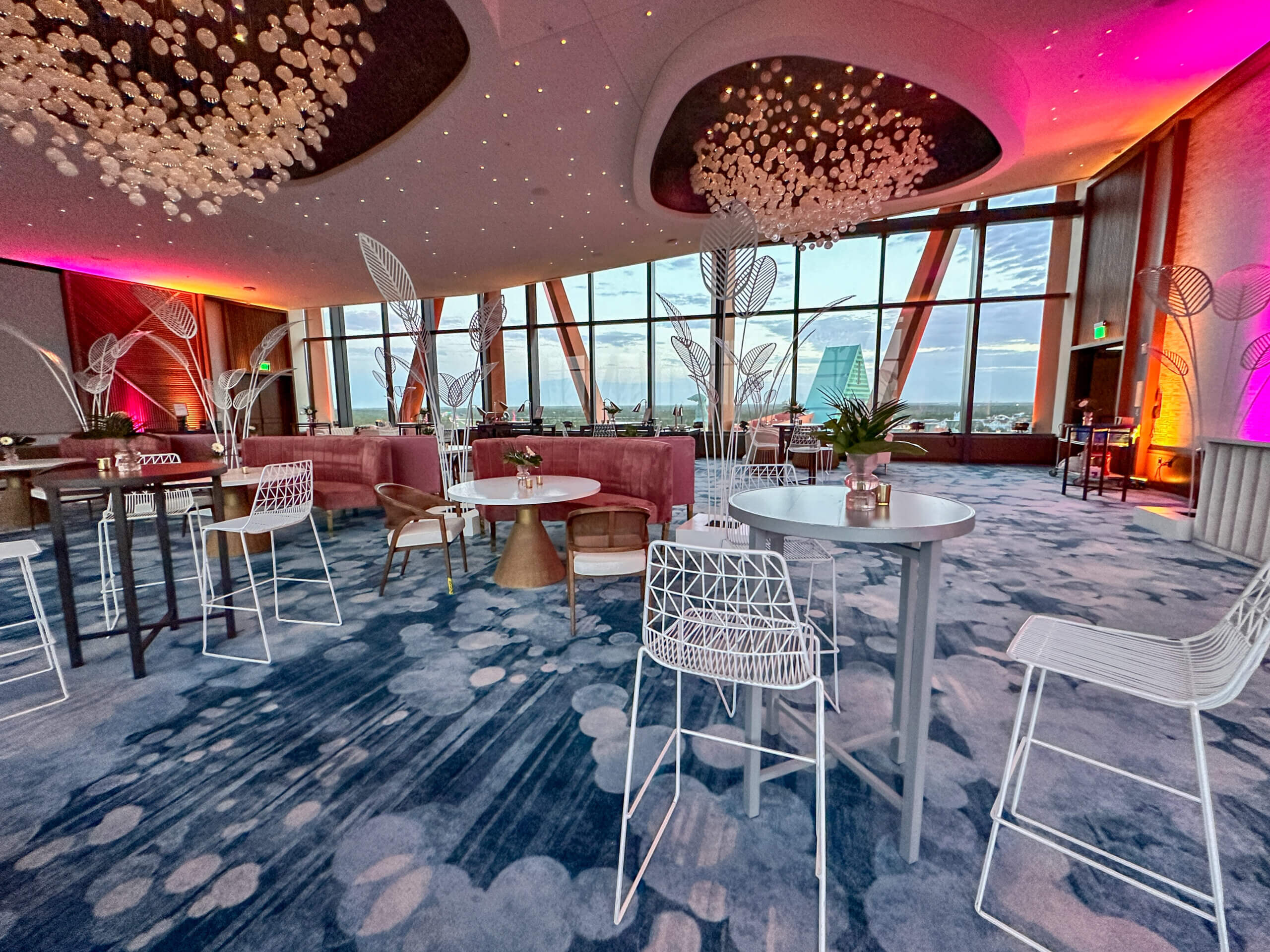 Современный и стильный ресторан « lounge » с ковровым покрытием с синим узором, белыми стульями и столами, а также большими окнами, из которых открывается вид на город. На потолке установлены художественные светильники, а стены освещаются фиолетовым и оранжевым светом.