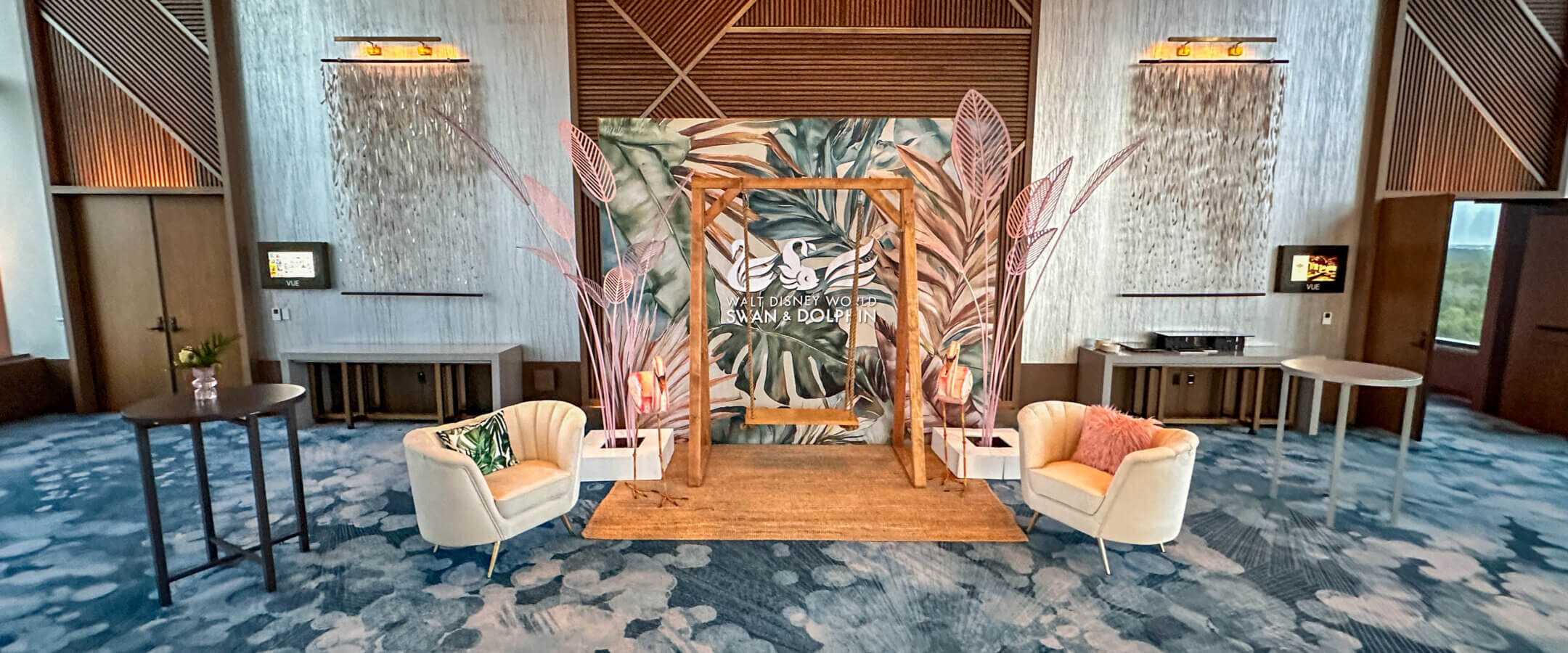 A modern event space with a decorative backdrop featuring leaves and Arabic script, flanked by white armchairs with colorful pillows, set on a blue patterned carpet and surrounded by wood accents and wall sconces.