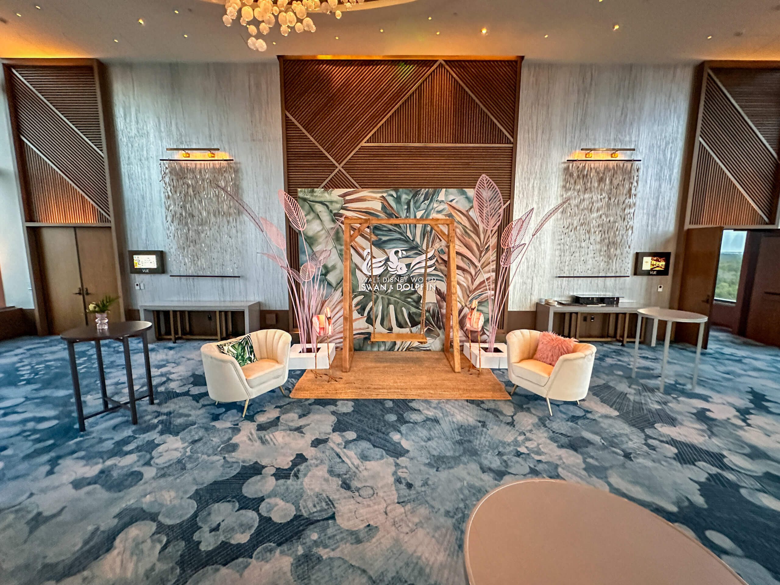 A modern event space with a decorative backdrop featuring leaves and Arabic script, flanked by white armchairs with colorful pillows, set on a blue patterned carpet and surrounded by wood accents and wall sconces.