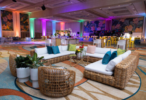 A spacious, colorful event hall with wicker sofas and chairs, white cushions, and vibrant pillows. Potted plants decorate the seating area, while colorful lights illuminate the walls and banquet tables are set up in the background.