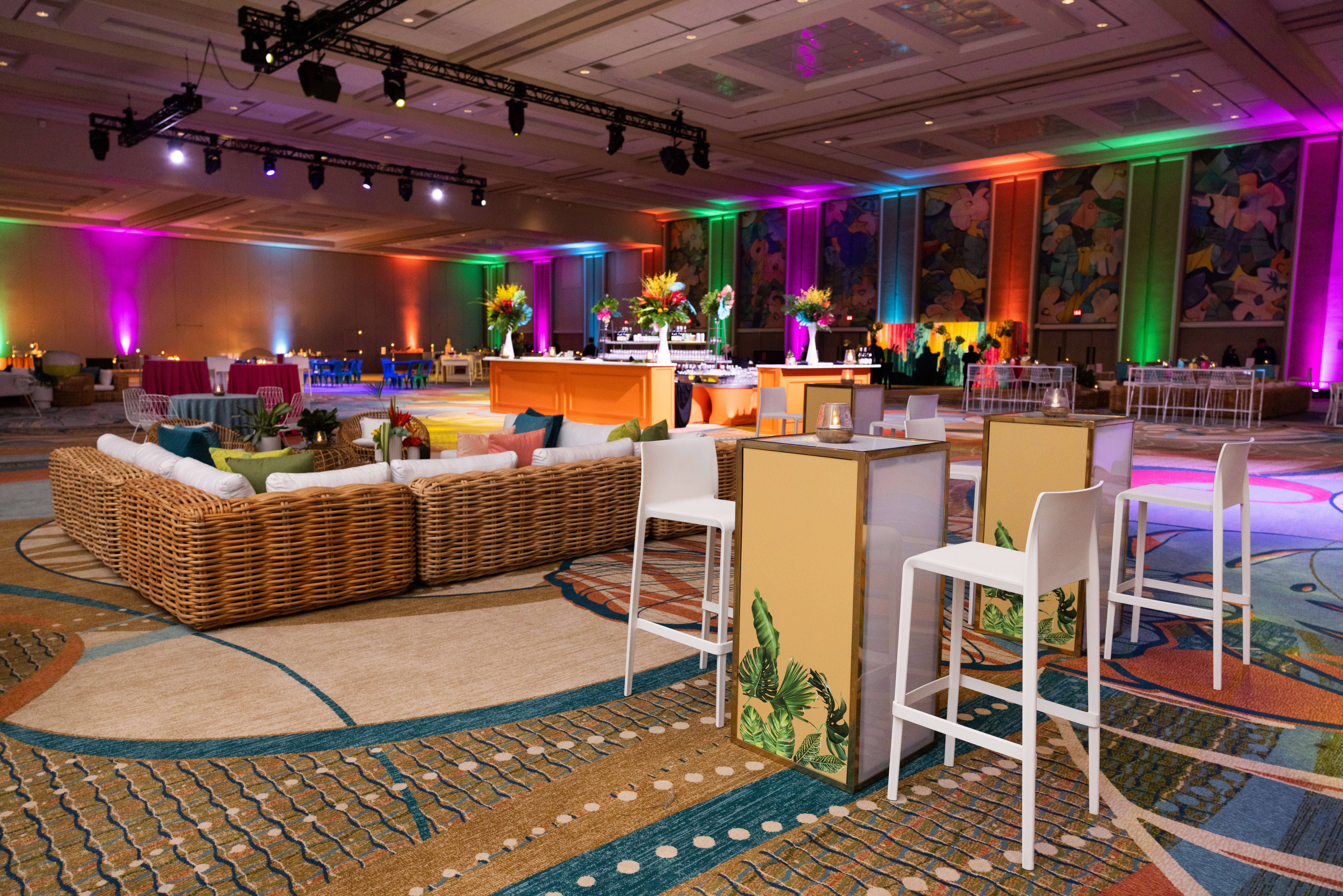 A spacious, colorful event hall with wicker couches, high tables and chairs, tropical-themed decor, buffet stations, and vibrant uplighting on the walls. The room has a festive, modern atmosphere.