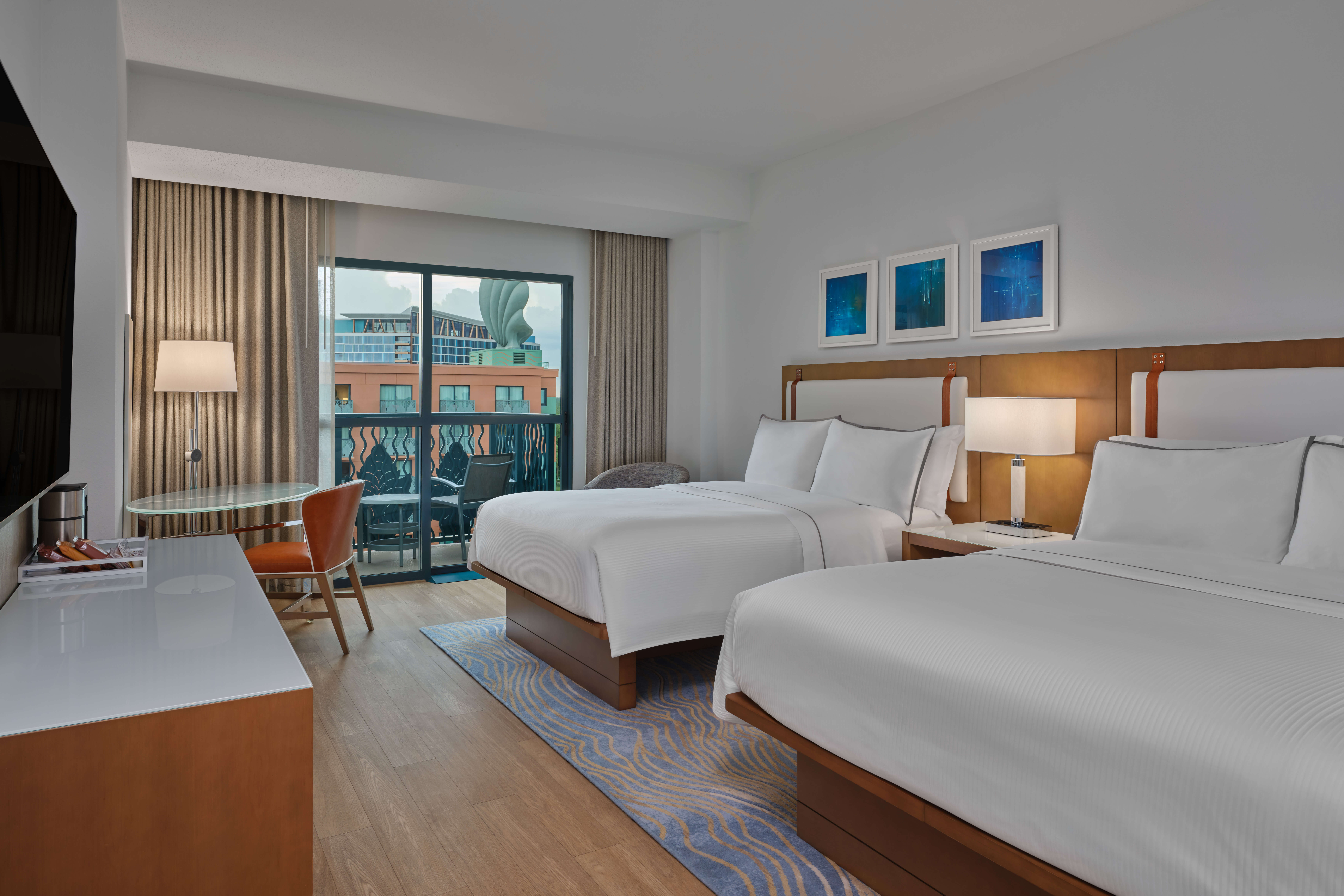 Modern hotel room with two double beds, white bedding, a desk and chair by a window, wall art above the beds, and a balcony with outdoor views. Soft lighting creates a comfortable and inviting atmosphere.