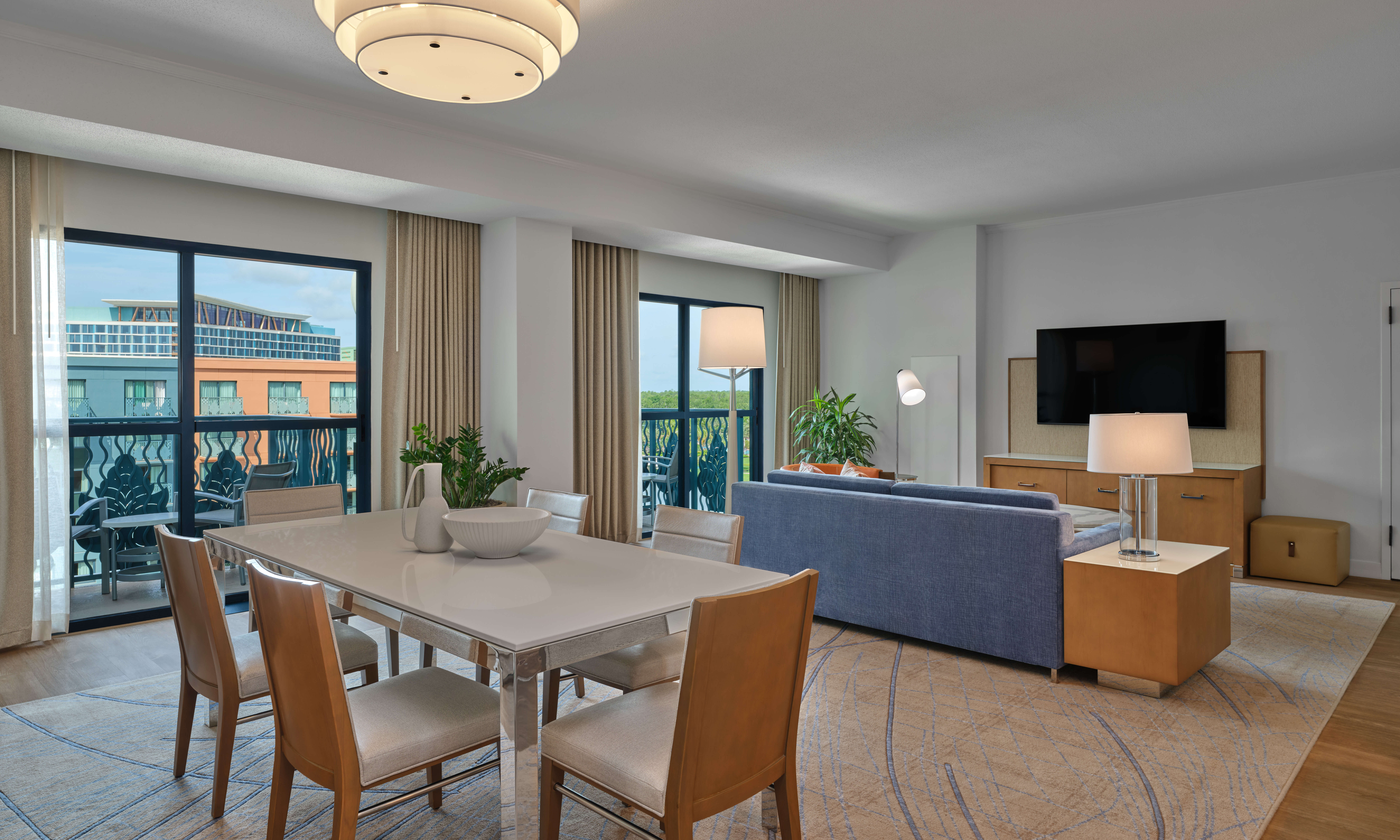 Modern living and dining area with a white table and six chairs, light blue sofa, TV, large windows with curtains, indoor plants, and balcony view of a building outside. Room is bright and well-lit.