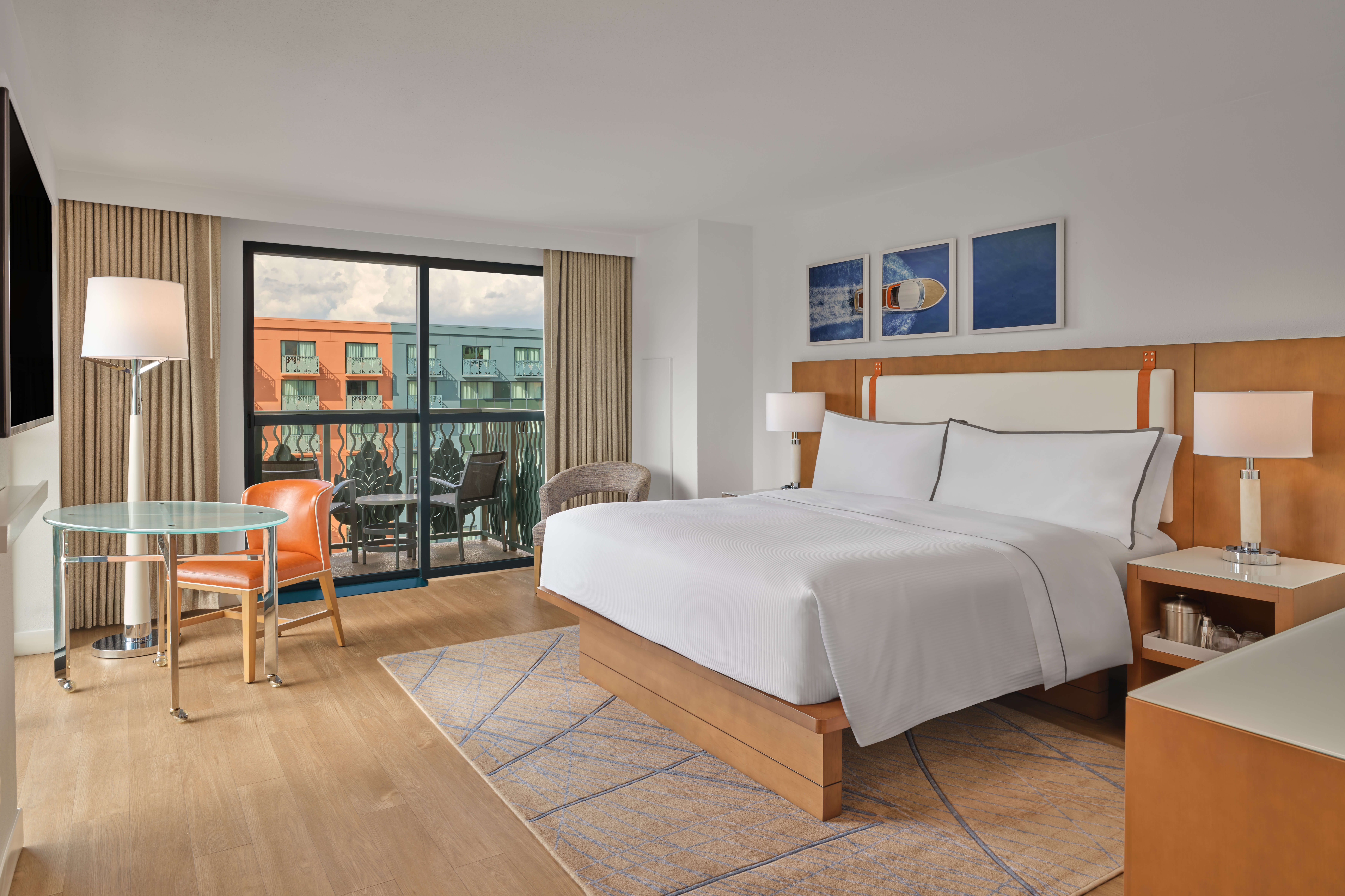 A modern hotel room with a large bed, glass desk and chair, two bedside tables with lamps, and a sliding glass door leading to a balcony with a view of colorful buildings outside.