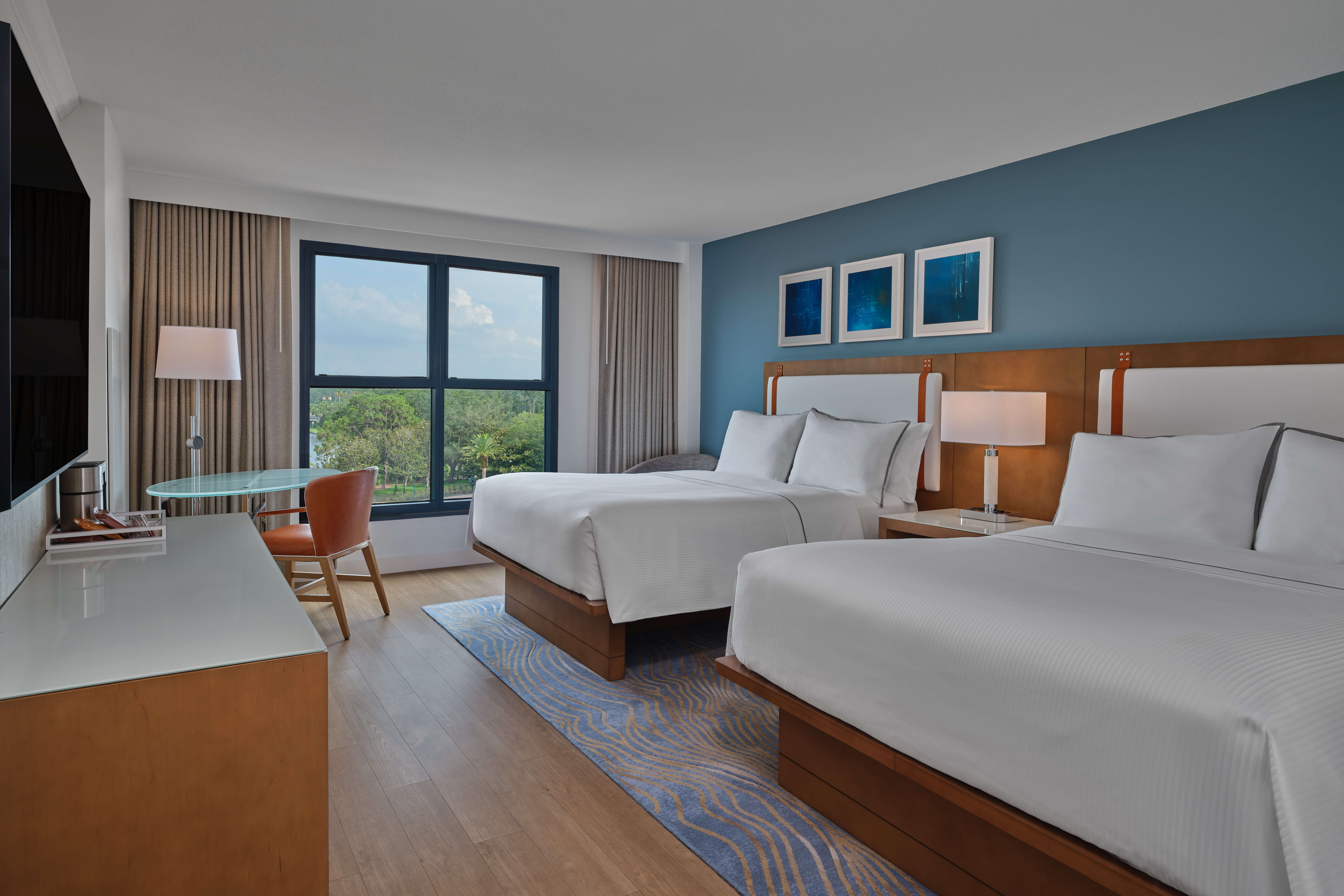 A modern hotel room with two double beds, white bedding, wood furniture, a blue accent wall, three framed art pieces, a desk with a chair, a floor lamp, and a large window with a view of trees and sky.
