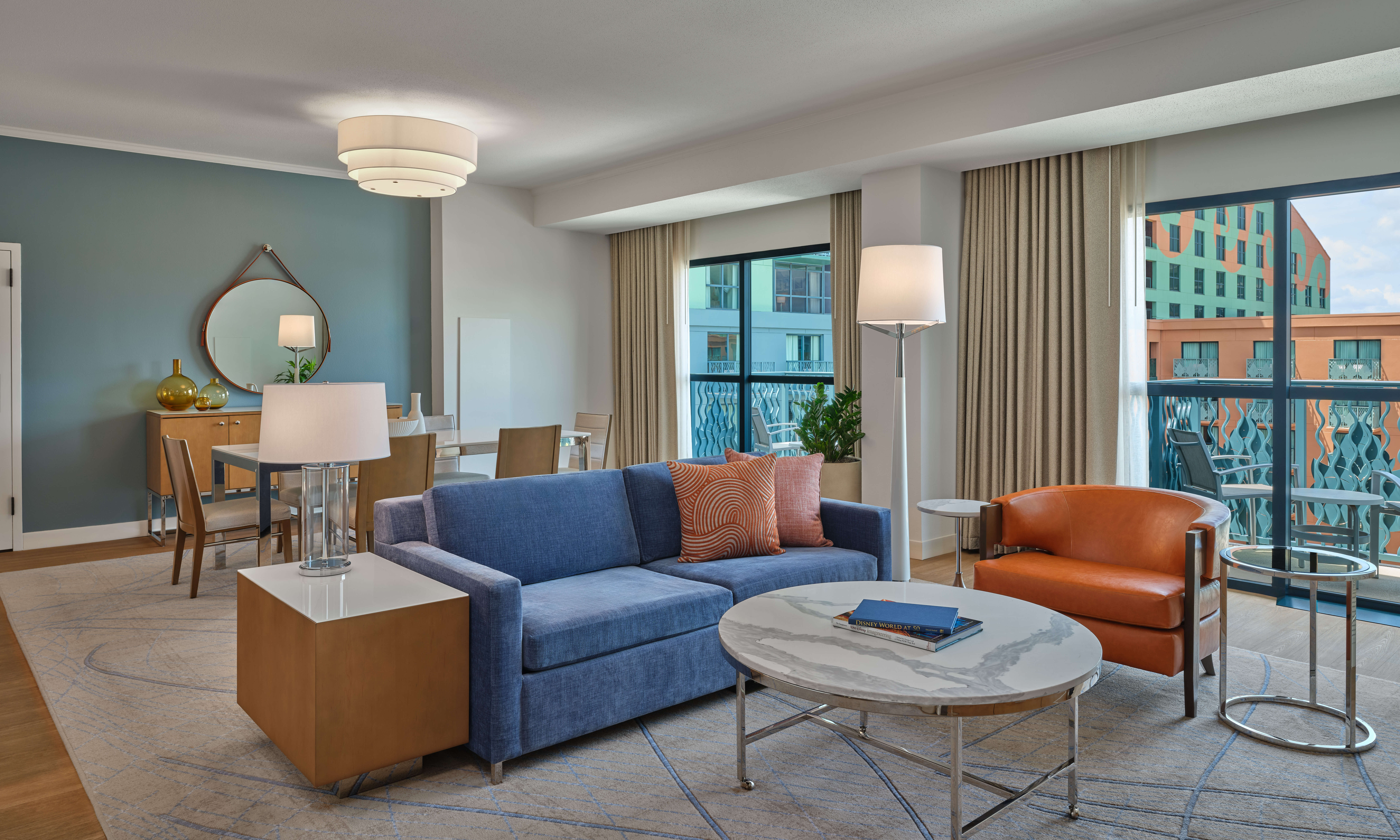 Modern hotel suite with a blue sofa, orange armchair, marble coffee table, and dining area. Large windows with curtains let in natural light and offer a view of colorful buildings outside.